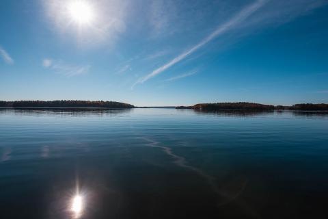 A photo of Great Bay, calm water reflects a blue sky with small streaks of clouds and a bright sun above a faraway tree-lined shoreline