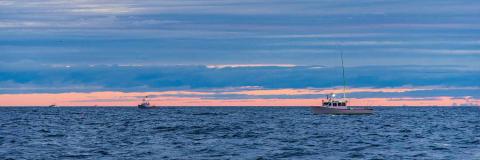 Two fishing boats float on a dark blue ocean under a sunrise with blue clouds