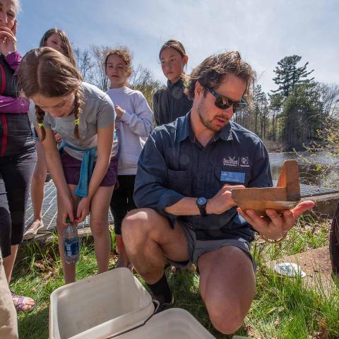 A CRV staff member wearing a blue shirt holds a fish measuring board as several children and people look on